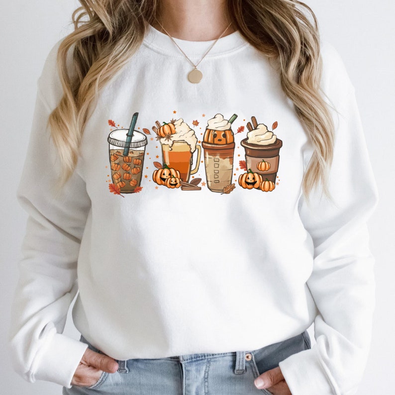 May include: White sweatshirt with a graphic of four fall-themed drinks with pumpkin faces. The drinks include a pumpkin spice latte, a pumpkin beer, a pumpkin milkshake, and a pumpkin iced coffee.