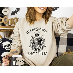 May include: Sand-colored sweatshirt with a black graphic of a skeleton reading a book and holding a drink. The text reads "I LIKE MY BOOKS SPICY & MY COFFEE ICY". The sweatshirt is a casual, comfortable style.