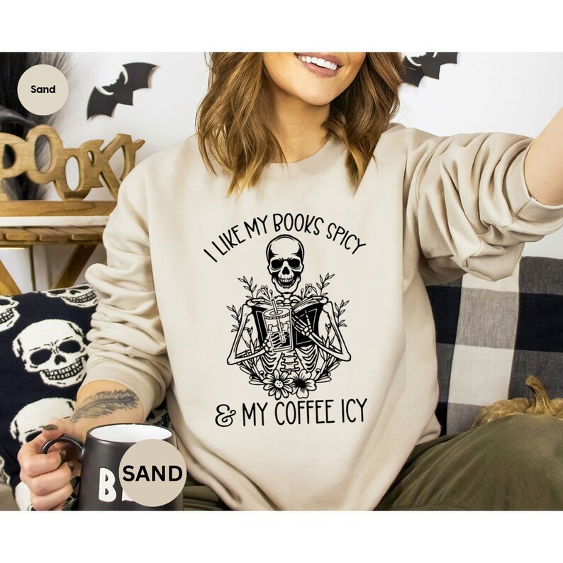 May include: Sand-colored sweatshirt with a black graphic of a skeleton reading a book and holding a drink. The text reads "I LIKE MY BOOKS SPICY & MY COFFEE ICY". The sweatshirt is a casual, comfortable style.