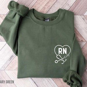 May include: Military green sweatshirt with a white heart-shaped design featuring "RN" and a stethoscope. The text "Evelyn" is also included. The sweatshirt is made of a soft material and has a crew neck.
