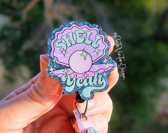 May include: A blue and pink glitter shell with a pearl and the text "Shell Yeah" on it. The shell is attached to a retractable badge holder.
