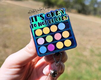 May include: A blue and green iridescent badge holder with a colorful eyeshadow palette design. The badge holder has a retractable clip and says "It's okay to not be okay".