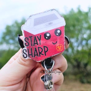 May include: A red and white glitter badge holder with a cartoon image of a biohazard container with the text "STAY SHARP". The badge holder has a black retractable clip.