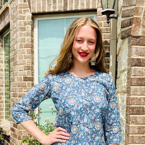 May include: A woman with long brown hair wearing a blue and white floral print top with a round neckline. She is standing in front of a brick wall with a window behind her.