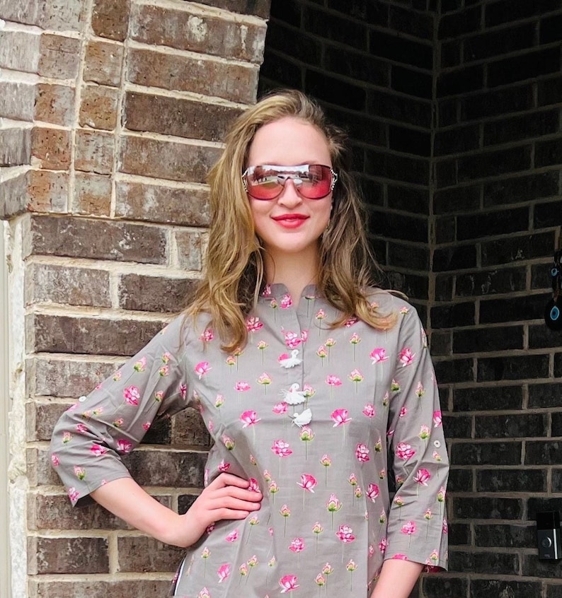 Peut inclure: Une femme portant un haut gris &agrave; imprim&eacute; floral avec des boutons blancs. Elle porte des lunettes de soleil roses et a la main sur la hanche.