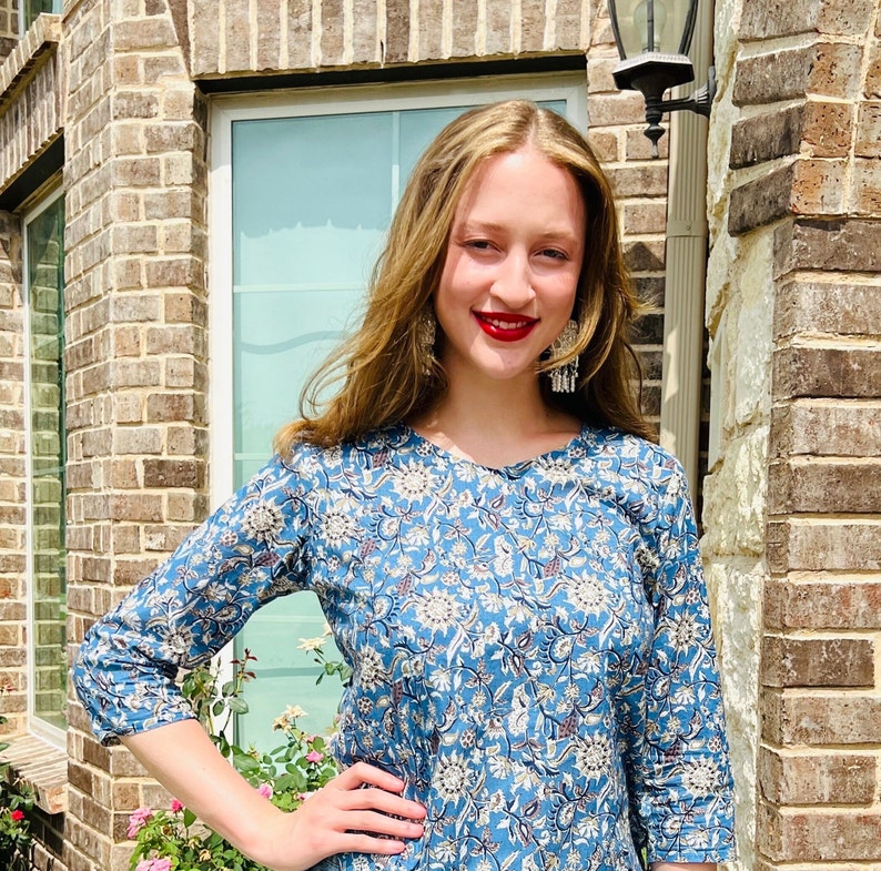 May include: A woman with long brown hair wearing a blue and white floral print top with a round neckline. She is standing in front of a brick wall with a window behind her.