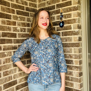 May include: A woman wearing a blue and white floral print top with 3/4 sleeves and blue jeans. She is standing in front of a brick wall with a blue evil eye charm hanging on it.