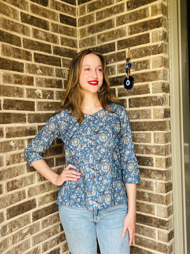 May include: A woman wearing a blue and white floral print top with 3/4 sleeves and blue jeans. She is standing in front of a brick wall with a blue evil eye charm hanging on it.