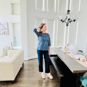 K&ouml;nnte beinhalten: Eine Frau tr&auml;gt ein blau-wei&szlig; gemustertes Oberteil, eine dunkelblaue Jeans und wei&szlig;e Plateau-Sandalen. Sie steht in einem Raum mit einem wei&szlig;en Sofa und einem Holztisch.