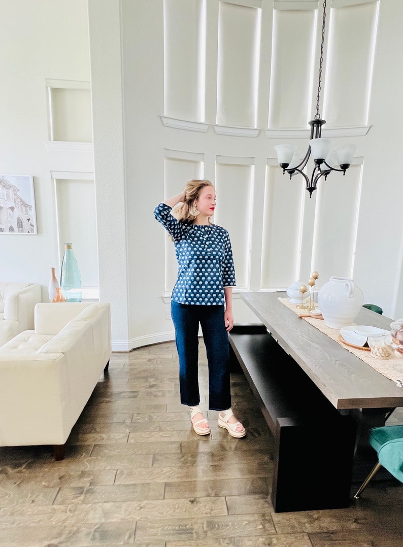 K&ouml;nnte beinhalten: Eine Frau tr&auml;gt ein blau-wei&szlig; gemustertes Oberteil, eine dunkelblaue Jeans und wei&szlig;e Plateau-Sandalen. Sie steht in einem Raum mit einem wei&szlig;en Sofa und einem Holztisch.