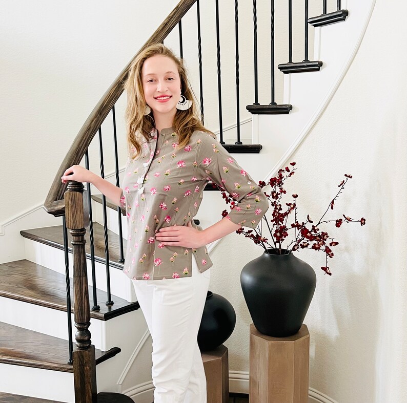 Peut inclure: Une femme portant un chemisier &agrave; fleurs gris avec un pantalon blanc se tient sur un escalier. Elle tient la rampe avec sa main gauche et sa main droite est sur sa hanche. Il y a un vase noir avec des fleurs rouges sur un pi&eacute;destal en bois en arri&egrave;re-plan.