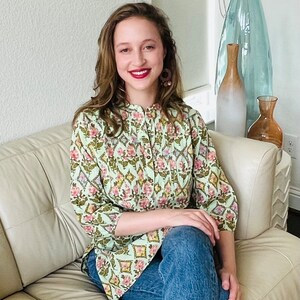 以下が含まれることがあります： 白い革のソファに座っている、ボタンダウンの襟と青いジーンズを履いた、薄い緑色の花柄のブラウスを着た女性です。