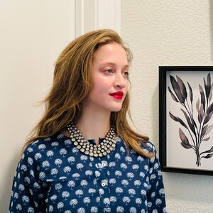 K&ouml;nnte beinhalten: Eine Frau mit langen braunen Haaren tr&auml;gt ein blau-wei&szlig; gemustertes Hemd mit einer mehrreihigen silbernen Halskette. Sie schaut nach rechts aus dem Bild.