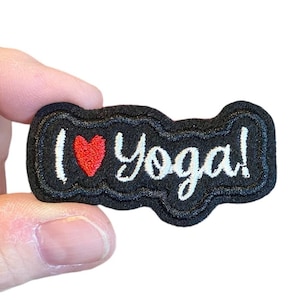 May include: A black patch with the phrase "I ❤️ Yoga!" embroidered in white and red. The patch has a wavy border and is held against a white background.