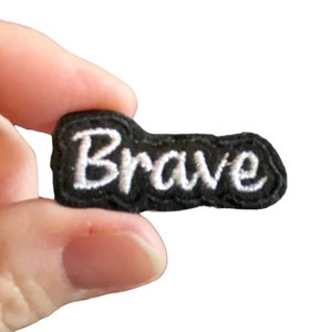 May include: Black fabric patch with white embroidered text that reads "Brave".