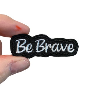May include: A black fabric patch with white embroidered text that reads "Be Brave".