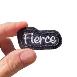 May include: A black embroidered patch with the word "Fierce" in white cursive lettering. The patch has a black border and is held by a hand against a white background. The patch is a small, rectangular shape.