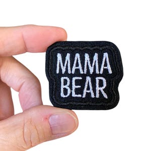 May include: A black embroidered patch with the words "MAMA BEAR" in white, block letters. The patch has a black border and is held against a white background.
