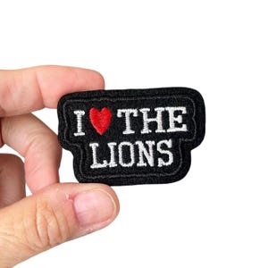 Op de afbeelding: Een zwart geborduurd embleem met de woorden "I ❤️ THE LIONS" in witte letters. Het hartsymbool is rood. De patch heeft een zwarte rand en is rechthoekig met afgeronde hoeken.