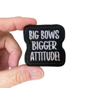 May include: Black embroidered iron-on patch with white text that reads "Big Bows Bigger Attitude!"