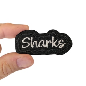 Może przedstawiać: Czarna naszywka haftowana z białym napisem "Sharks".