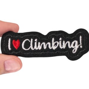 Puede incluir: Un parche bordado negro con la frase "I ❤️ Climbing!" en escritura blanca, con un corazón rojo. El parche tiene un borde ondulado y está sostenido por una mano sobre un fondo blanco. El parche es un accesorio temático de escalada.