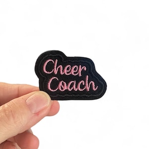 May include: Black iron-on patch with pink embroidered text that reads "Cheer Coach".