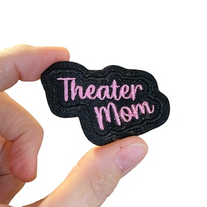 May include: A black embroidered patch with the words "Theater Mom" in pink script. The patch has a cloud-like shape with a textured border. The patch is held between two fingers against a white background.