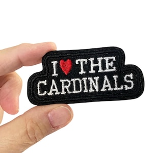 Puede incluir: Parche bordado negro con las palabras "I ❤️ THE CARDINALS" en letras blancas. Un corazón rojo reemplaza la palabra "love". El parche tiene un borde negro y está sostenido por una mano.