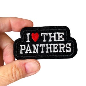 Op de afbeelding: Zwart geborduurde patch met de tekst "I ❤️ THE PANTHERS" in witte letters. Een rood hart vervangt het woord "love". De patch heeft een zwarte rand en wordt door een hand vastgehouden.
