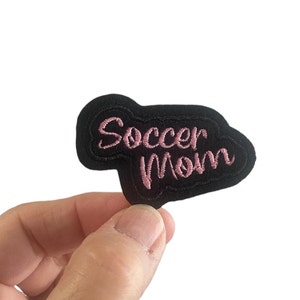 May include: Black felt patch with pink embroidered text that reads "Soccer Mom".