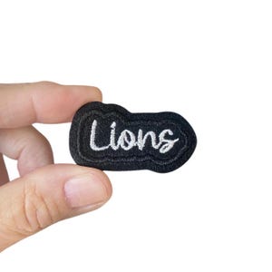 May include: Black felt patch with white embroidered text that reads "Lions".