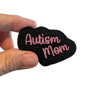 May include: A black embroidered patch with the words "Autism Mom" in pink script. The patch has a unique, cloud-like shape and is likely designed to be sewn or ironed onto fabric items.