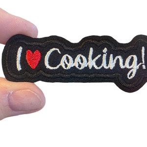 Può includere: Toppa ricamata nera con testo bianco che dice "Amo cucinare!" con un cuore rosso.