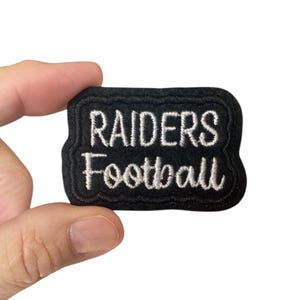 Puede incluir: Parche bordado negro con texto blanco que dice "RAIDERS Football".