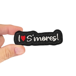 May include: Black embroidered patch with the text "I ♥ S'mores!" in white script, a red heart symbol, and a black border. The patch is held against a white background.