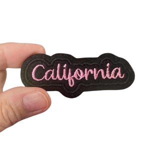 Puede incluir: Parche de planchado negro con texto bordado en rosa que dice "California".