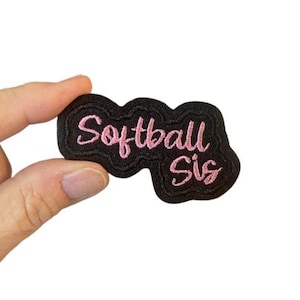 Peut inclure: Écusson brodé noir avec les mots "Softball Sis" en écriture rose. Le patch a une bordure ondulée. Le patch est tenu par une main sur fond blanc.