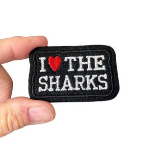 May include: Black rectangular patch with the phrase "I ❤️ THE SHARKS" embroidered in white thread. A red heart replaces the word "love". The patch has a black border.