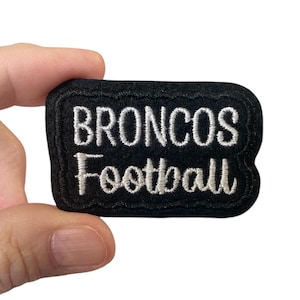 May include: A black rectangular patch with white embroidered text reading "BRONCOS Football". The patch has a black border and is held by a hand.