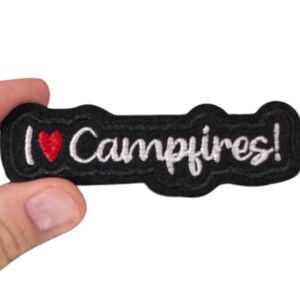 May include: A black patch with the phrase "I ❤️ Campfires!" embroidered in white script. The heart is red. The patch is held by a hand.