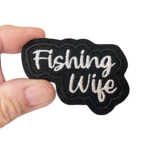 Puede incluir: Un parche bordado negro con las palabras "Fishing Wife" en escritura blanca. El parche tiene un diseño texturizado en capas y está sostenido por una mano sobre un fondo blanco. Este parche es un accesorio divertido para los entusiastas de la pesca.