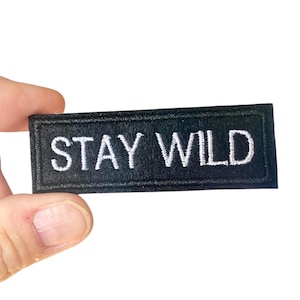 May include: A black rectangular patch with the words "STAY WILD" embroidered in white. The patch is held against a white background, showcasing the contrasting colours and the clear, bold text.