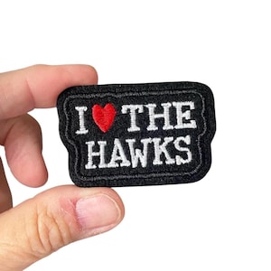 Könnte beinhalten: Schwarzer, gestickter Aufnäher mit dem Text "I ❤️ THE HAWKS" in weißen Buchstaben und einem roten Herzsymbol. Der Aufnäher hat einen schwarzen Rand und ist rechteckig.