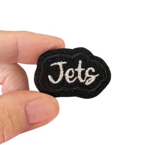 May include: A black felt patch with white embroidered lettering that reads "Jets".