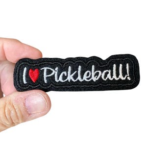 May include: Black embroidered patch with the words "I ❤️ Pickleball!" in white script and a red heart. The patch has a wavy border. The patch is held against a white background.
