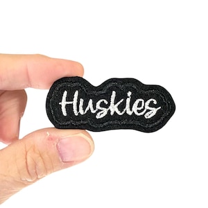 May include: Black embroidered patch with the word "Huskies" in white script. The patch has a wavy border and is held between fingers against a white background. Ideal for jackets, bags, or other fabric items.