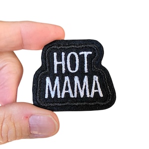 May include: A black embroidered patch with the words "HOT MAMA" in white, stylized lettering. The patch has a rounded rectangular shape and is held against a white background.