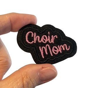 May include: A black embroidered patch in a cloud shape with the words "Choir Mom" in pink script. The patch has a black border and is held by a hand against a white background.