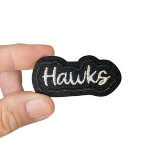 Könnte beinhalten: Schwarzer gestickter Aufnäher mit weißer Aufschrift "Hawks".